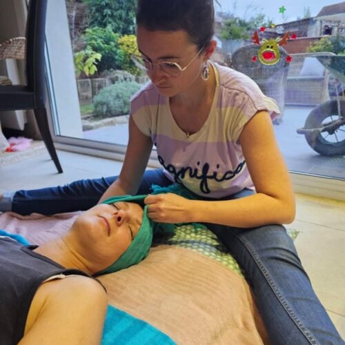 femme assise avec un tee shirt manches courtes rayé beige et violet avec écrit magnifico. Elle est brune les cheveux attachés, elle porte des lunettes. Elle porte un jean. Elle se tient assise les jambes écartées, elle tient un tissu bleu, qu'elle semble manipuler, la tete d'un autre femme y est déposée, elle a les yeux fermés et le visage sur le côté déposé dans le tissu. La femme assise tient le tissu d'une main et son autre main semble être sous la tête. Le tissu fait une sorte de bandeau sur la tete de l'autre femme. Sous ses épaules on peut voir un autre tissu bleu. Elle est allongée sur un matelas au sol et elle porte un débardeur noir, on ne voit que le haut de son corps. Autour, on peut voir une chaise, une baie vtrée avec décoration dessus et la vue sur un jardin.