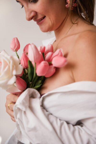 femme avec chemisier blanc. Tiens des fleurs entre ses mains. Se tient debout de profil avec une épaule dégagée. Porte une boucle d'oreille fleur rose et une bague couleur or. Elle sourit et regarde le bouquet.