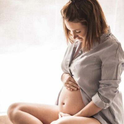 femme enceinte de profil, assise sur un coussin. Elle porte une chemise ouverte au niveau de son ventre. Elle a une main posée sur le haut de son ventre et une en dessous. Elle a les jambes dégarnies. Ses cheveux sont détachés au carré. Il y a un joli voilage blanc en arrière plan