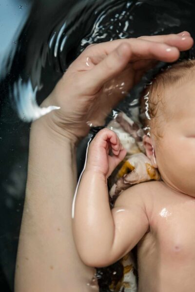 photo d'un bébé dans un bain. on peut voir une partie de son corps, sa main et son bras droit. On peut voir une partie du lange. Il y a également une main et un avant bras d'adulte à côté. La main semble vouloir caresser la tête. On peut voir l'onde dans l'eau.