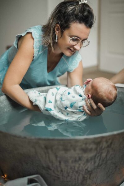 baignoire en zinc dans laquelle une femme fait le bain enveloppé d'un bébé. Elle est face à lui, elle porte un haut bleu à manches courtes avec des petites fleurs blanches sur les bords. Elle porte des boucles d'oreilles blanches, des lunettes, elle est brune les cheveux détachés au carré. On ne voit pas le visage du bébé. Il est emmailloté dans un lange blanc avec des petits motifs bleus.