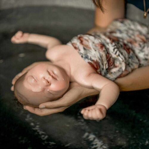 baingoire en zinc dans laquelle un bébé est baigné. il est endormi, il a un oeil dans l'eau et le reste de son corps semble détendu. ses bras sont ouverts; Il est dans un lange fleuri. Il est tenu dans les mains d'une autre personne.