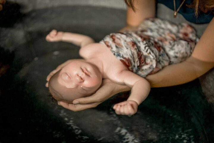 baignoire en zinc dans laquelle un bébé est baigné. il est endormi, il a un oeil dans l'eau et le reste de son corps semble détendu. ses bras sont ouverts; Il est dans un lange fleuri. Il est tenu dans les mains d'une autre personne.