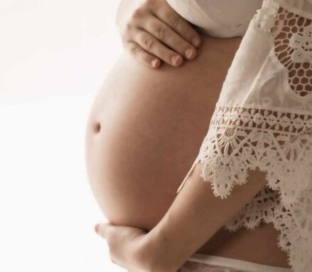 photo d'un ventre de femme enceinte. Zoom sur le ventre, on voit peu le reste du corps.une main est au dessus et l'autre en dessous. Elle porte un kimono blanc avec de la dentelle, il est ouvert. on peut apercevoir les sous vetements blancs.