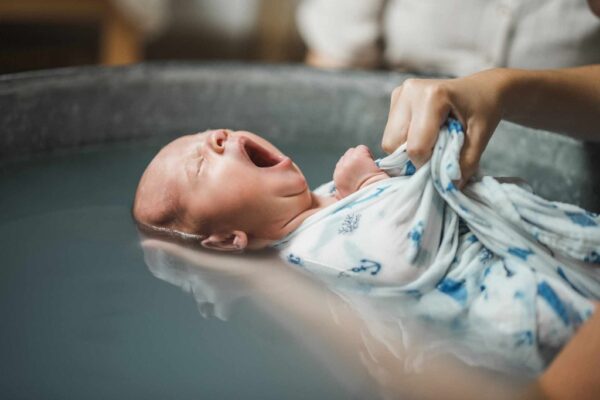 baignoire en zinc dans laquelle est baigné un bébé. Il baille, il est emmailloté dans un tissu blanc avec déssins dessus sur le thème marin : ancre, algues... il est tenu par une main qui soutient la tête et l'autre qui maintient le tissu.
