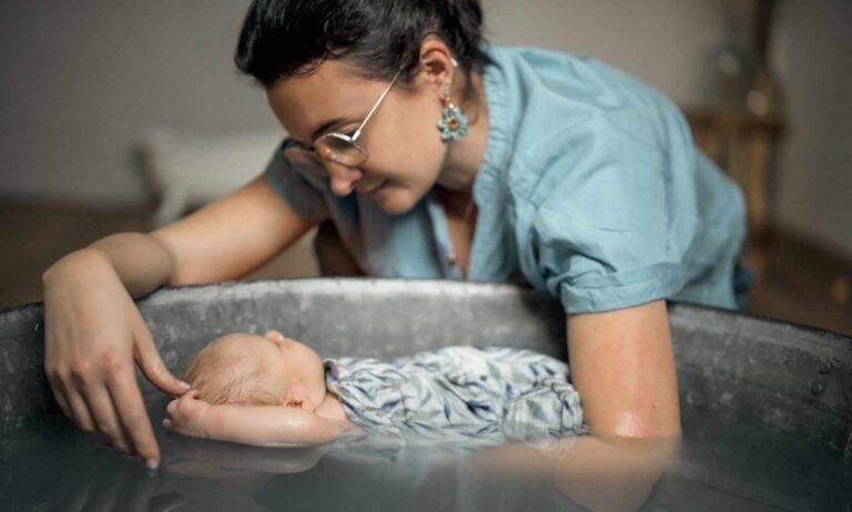 baignoire en zinc dans laquelle est réalisé le bain d'un bébé emmailloté dans un tissu fleuri dans les tons blanc, bleu, vert. son petit bras droit et sorti et il le met au dessus de sa tête. Le bain est donné par une jeune femme avec un haut bleu et des boucles d'oreille en forme de fleurs. Elle porte des lunettes. elle est brune et ses cheveux sont attachés. ils semblent se regarder tous les deux.