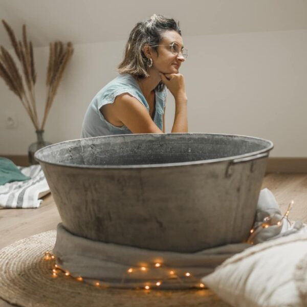 Photo avec en arrière plan de la pampa sur la gauche dans un vase. On peut voir une femme assise au sol, elle a les cheveux au carré détachés, elle porte des lunettes, des boucles d'oreille et un haut bleu à manches courtes. Elle est de profil le bras plié et la main sous le menton. au premier plan on peut voir une bassine en zinc avec une guirlande lumineuse autour au sol. elle est posée sur un tapis en jute et il y a un voilage autour de la bassine.