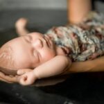 Bain d'un bébé emmailloté dans un lange. Ses bras son sortis. Ce bébé est endormi dans les bras de la personne qui le baigne, il semble si détendu. son petit bras droit et levé au dessus de sa tête. on ne voit que le bébé sur cette photo.