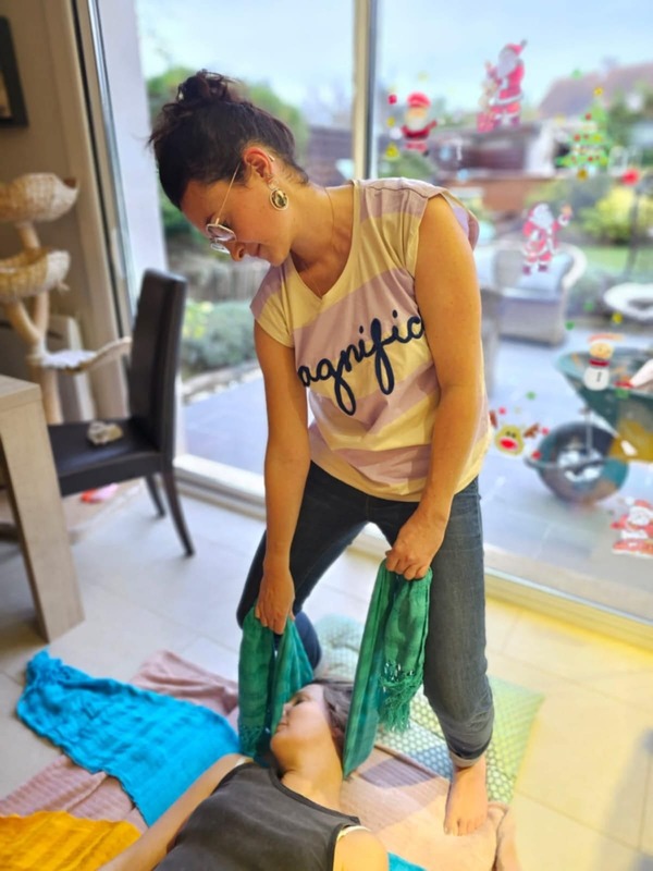 femme debout avec un tee shirt manches courtes rayé beige et violet avec écrit magnifico. Elle est brune les cheveux attachés, elle porte des lunettes. Elle porte un jean. Elle se tient debout les jambes fléchies, elle tient un tissu bleu, qu'elle semble manipuler, la tete d'un autre femme y est déposée, elle a les yeux fermés et le visage sur le côté déposé dans le tissu. Sous ses épaules on peut voir un autre tissu bleu et plus bas un tissu jaune. elle est allongé sur un matelas au sol et elle porte un débardeur noir, on ne voit que le haut de osn corps. autour on peut voir une chaise, une baie vtrée avec décoration dessus et la vue sur un jardin.