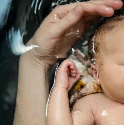 photo d'un bébé dans un bain. on peut voir une partie de son corps, sa main et son bras droit. On peut voir une partie du lange. Il y a également une main et un avant bras d'adulte à côté. La main semble vouloir caresser la tête. On peut voir l'onde dans l'eau.