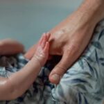 bébé dans un lange, les bras sortis. Le lange est avec des fleurs bleues. On voit ses petits bras et ses petites mains mais pas son visage. Une des petites mains tient la main d'un adulte.