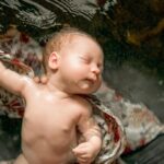 bain bébé, il est détendu, les yeux fermés dans les mains de quelqu'un. un lange à fleurs et sous son corps. On voit le haut de son corps avec sa petite main et son bras droit allongé à la perpendiculaire. son visage est de profil