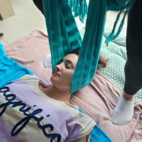 femme allongée sur un matelas au sol. on voit son buste et sa tête, elle a les yeux fermés. elle porte un tee shirt manches courtes rayés violet et beige écrit magnifico dessus. sa tête est dans un tissu bleu ciel soutenu par une autre personne par les deux extrémités. un autre tissu bleu est sous ses épaules
