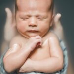 photo d'un bébé endormi lors d'un bain. Il est dans un lange blanc à fleurs bleues et grises. Ses bras et son torse sont en dehors du lange, il dort, son bras gauche est plié sur lui et son autre plié avec la main sous le menton. On peut voir de chaque partie de son visage les mains de celui qui le baigne