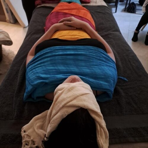 femme allongée les mains sur son ventre. Le haut de son visage est enveloppé dans un tissu beige, ses épaules dans un tissu bleu, elle porte un haut noir que l'on peut distinguer au niveau de sa poitrine, on voit un tissu jaune qui enveloppe son bassin et on aperçoit ensuite un autre tissu orange ou rouge. La photo de montre pas plus bas que le ventre.