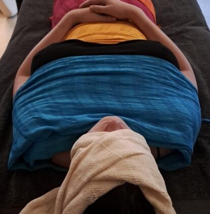 femme allongée les mains sur son ventre. Le haut de son visage est enveloppé dans un tissu beige, ses épaules dans un tissu bleu, elle porte un haut noir que l'on peut distinguer au niveau de sa poitrine, on voit un tissu jaune qui enveloppe son bassin et on aperçoit ensuite un autre tissu orange ou rouge. La photo de montre pas plus bas que le ventre.