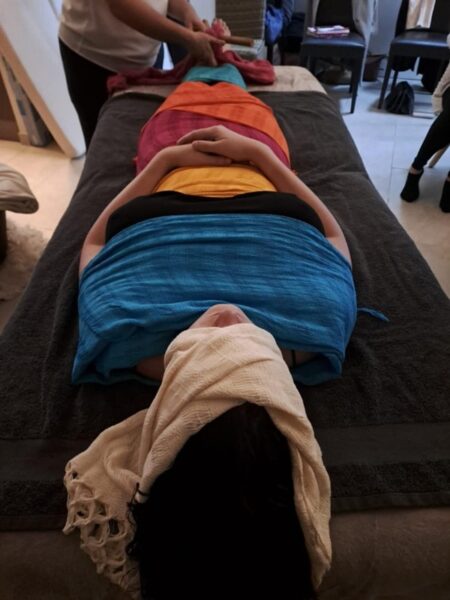 femme allongée les mains sur son ventre. Le haut de son visage est enveloppé dans un tissu beige, ses épaules dans un tissu bleu, elle porte un haut noir que l'on peut distinguer au niveau de sa poitrine, on voit un tissu jaune qui enveloppe son bassin et on aperçoit ensuite un autre tissu orange ou rouge. La photo de montre pas plus bas que le ventre.