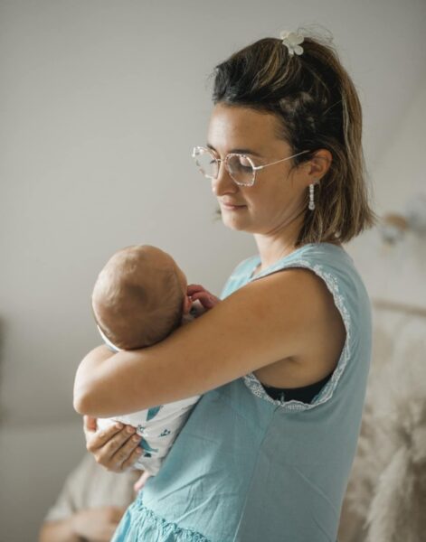 photo d'une femme debout, brune les cheveux au carré avec une pince fleur. elle porte des lunettes et des boucles d'oreilles blanches. Elle est de 3/4 , elle sourit en regardant le bébé qu'elle tient dans ses bras. Elle porte un haut à manches courtes bleu ciel. Le bébé est dans un tissu blanc à motif.