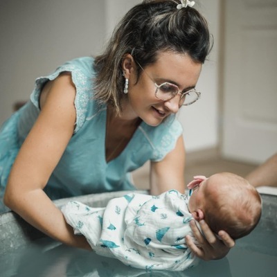 baignoire en zinc dans laquelle une femme fait le bain enveloppé d'un bébé. Elle est face à lui, elle porte un haut bleu à manches courtes avec des petites fleurs blanches sur les bords. Elle porte des boucles d'oreilles blanches, des lunettes, elle est brune les cheveux détachés au carré. On ne voit pas le visage du bébé. Il est emmailloté dans un lange blanc avec des petits motifs bleus. Elle sourit, elle est penchée au dessus de la bassine